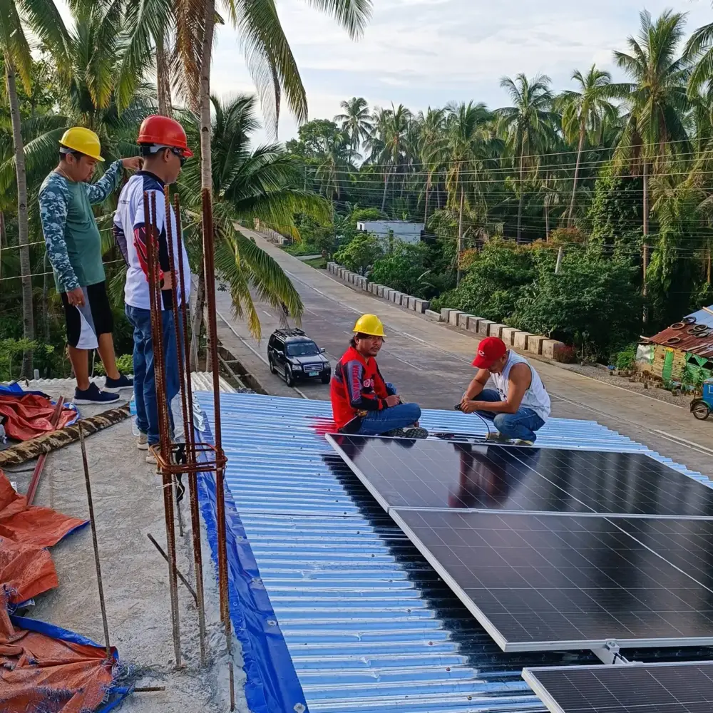 MACC crew mounting solar panels on a metal roof, with palm trees and a provincial road behind them
